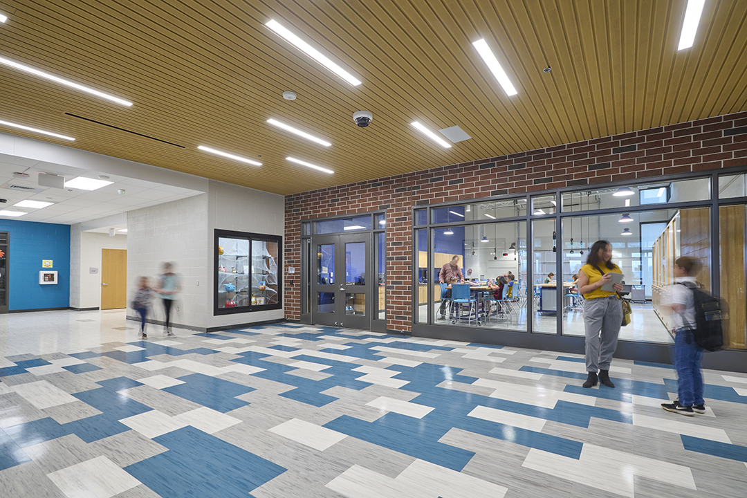Rossmoyne Elementary School Entry. Large wind permit students and visitors to look into a STEAM classroom. A student and teacher are standing in front of the windows talking.
