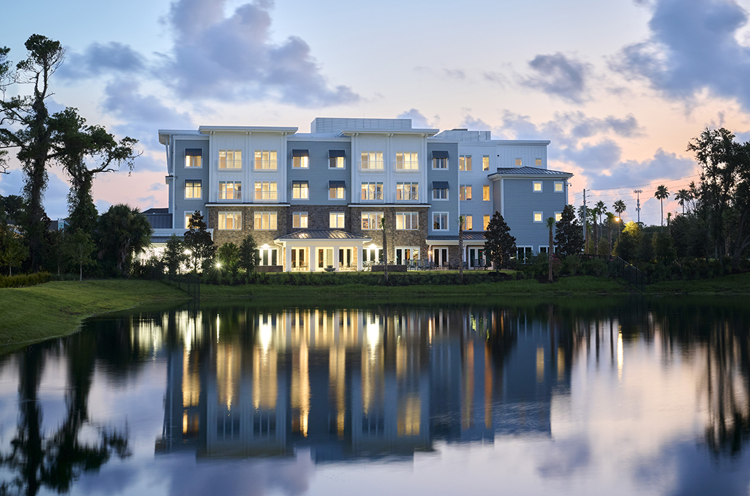 Exterior view of 4-story assisted living building at dusk with windows lit and building reflected in pond