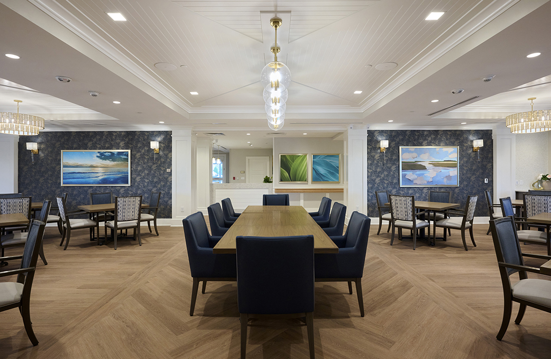 Assisted living dining room space with community table centered under accent lighting with local nature-inspired art
