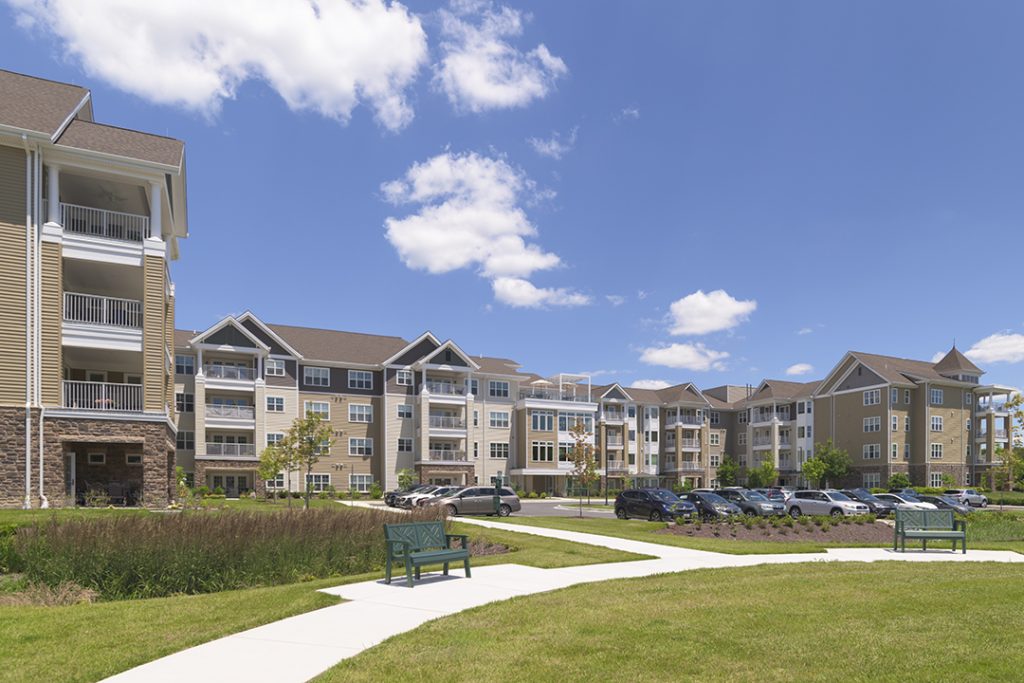 Four-story independent living apartment buildings surrounding landscaped walking paths, open green space, and resident parking.